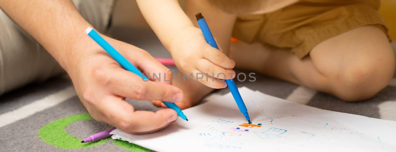 Little toddler boy of two or three years old with dad draws with markers in the album in the children's room at home sitting on the floor. Spending time with children. Selective focus
