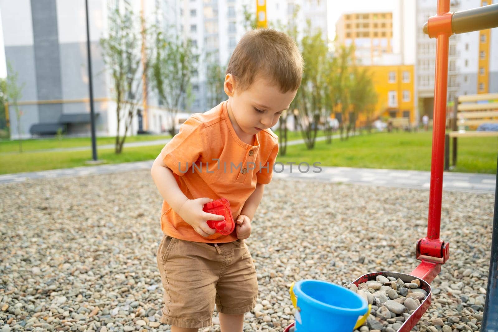 Little toddler boy playing with toys on pebbles on the playground. Boy two or three years old. Toddlerhood childhood concept. Residential complex of apartment buildings.