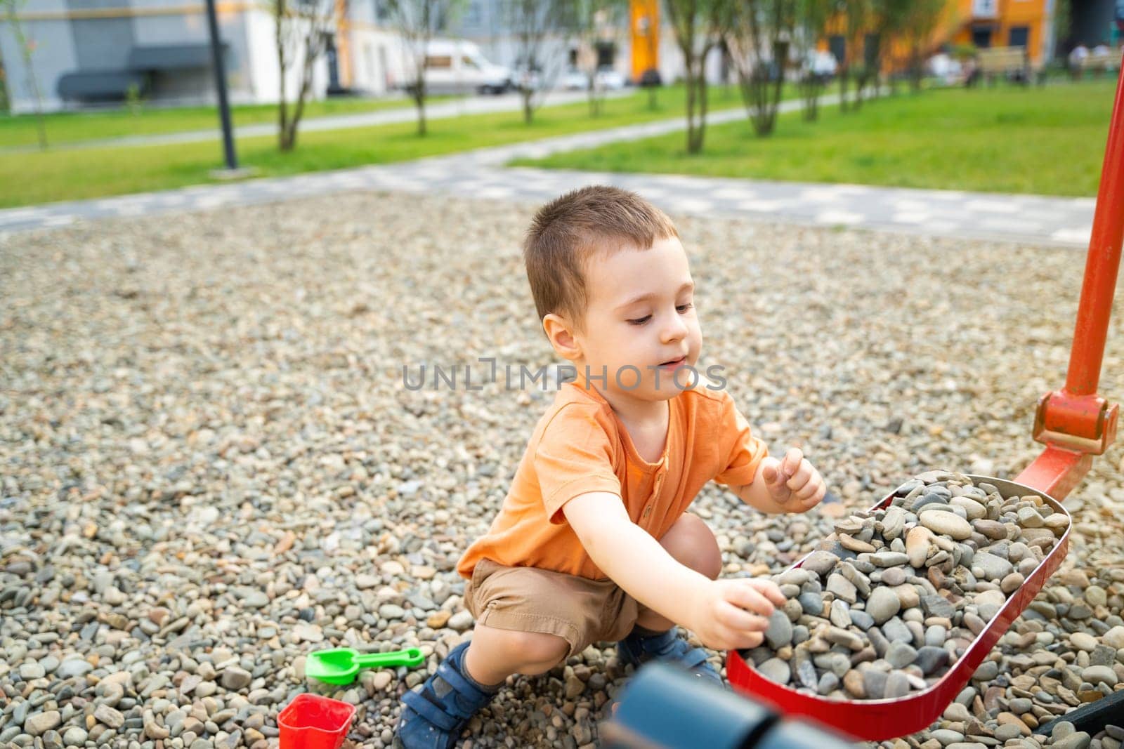 Little toddler boy playing with toys on pebbles on the playground. Boy two or three years old. Toddlerhood childhood concept. Residential complex of apartment buildings.