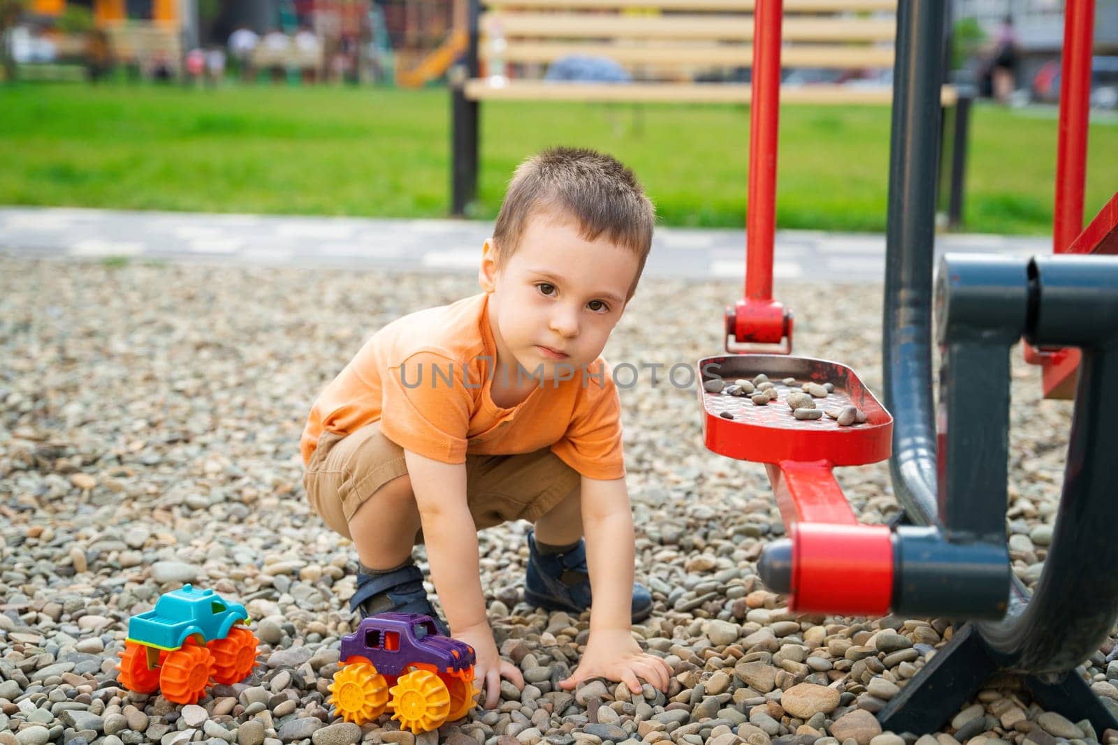 Little toddler boy playing with cars on pebbles on the playground. Boy two or three years old. Toddlerhood childhood concept. Residential complex of apartment buildings.