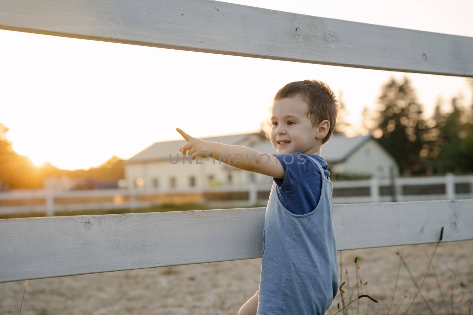 Happy toddler boy two or three years old stands near a fence and points with his finger on a sunny summer day. Summer sunset light. Horse farm