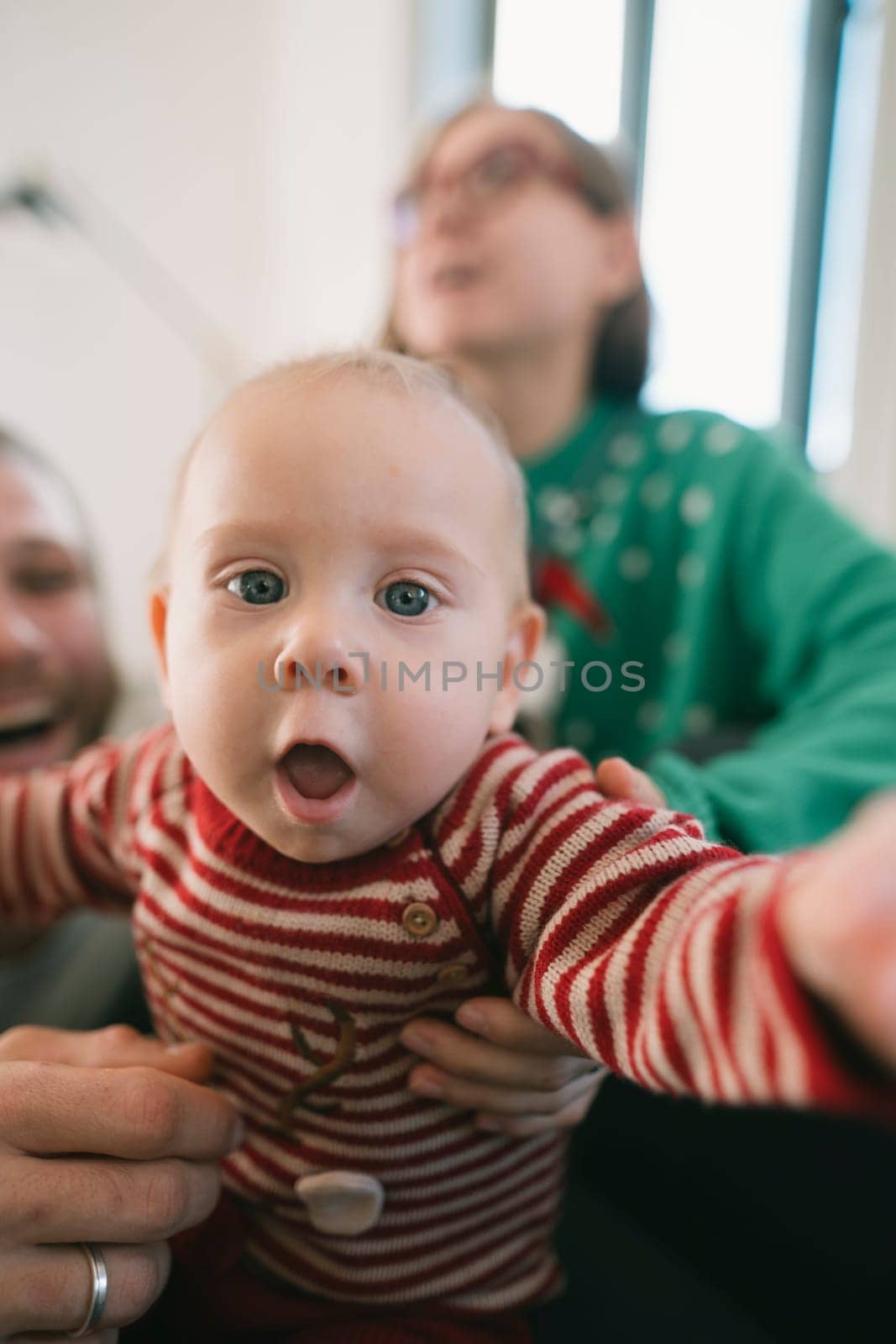 In a cozy setting, a playful baby reaches out to the camera, surrounded by loving, smiling family members