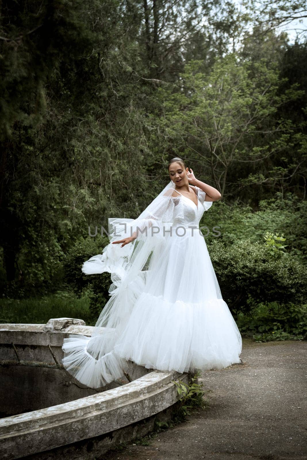 beautiful bride with long veil against the background of trees and old fountain. brunette girl in long white dress, she has smooth hair, old ruined fountain behind. veil reaches the ground