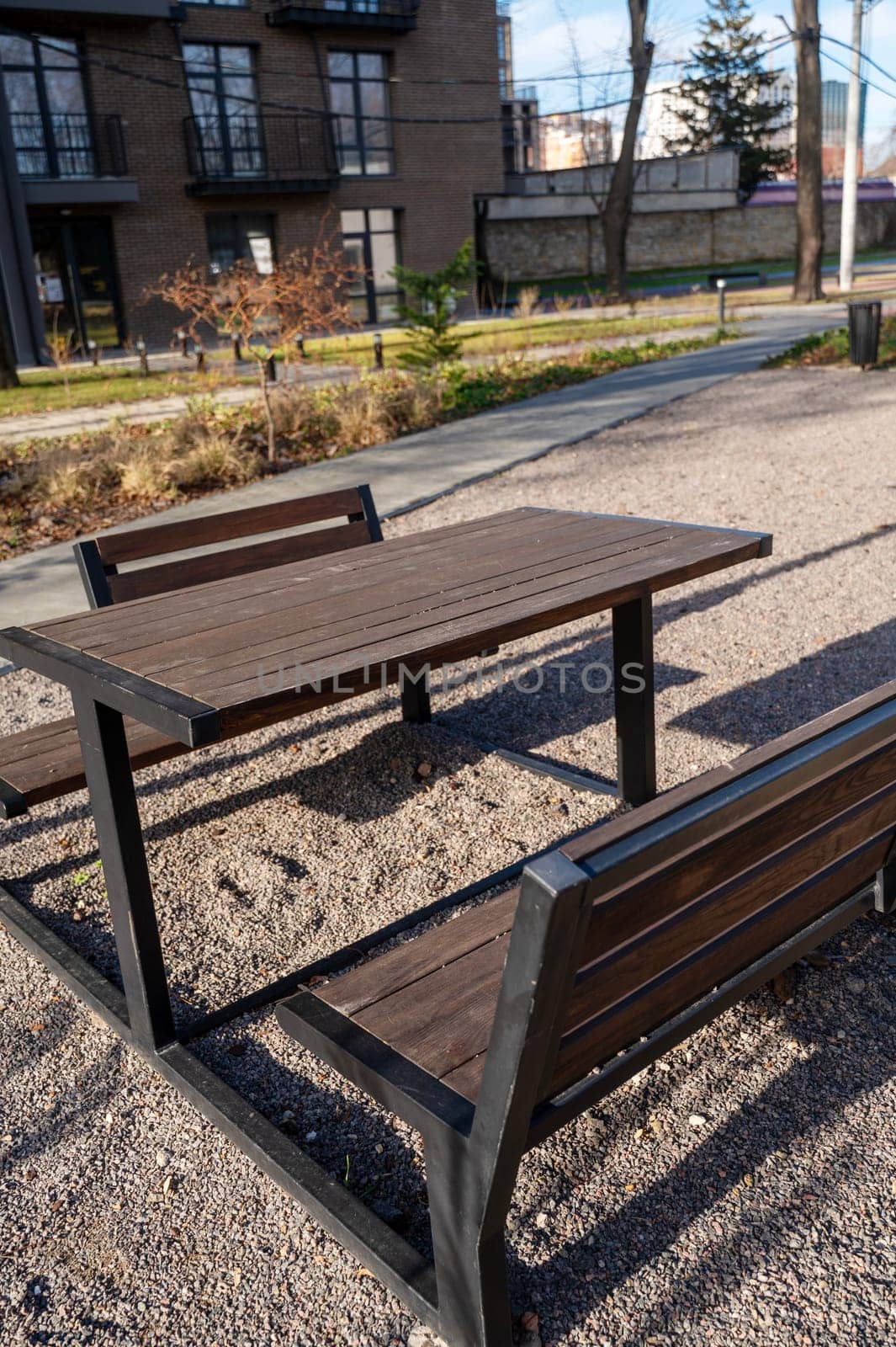 a large table and benches made of wood. benches and table are dark brown, stand in a recreation area in nature. on the ground small stones. around is clean, nearby is a pedestrian path made of tiles