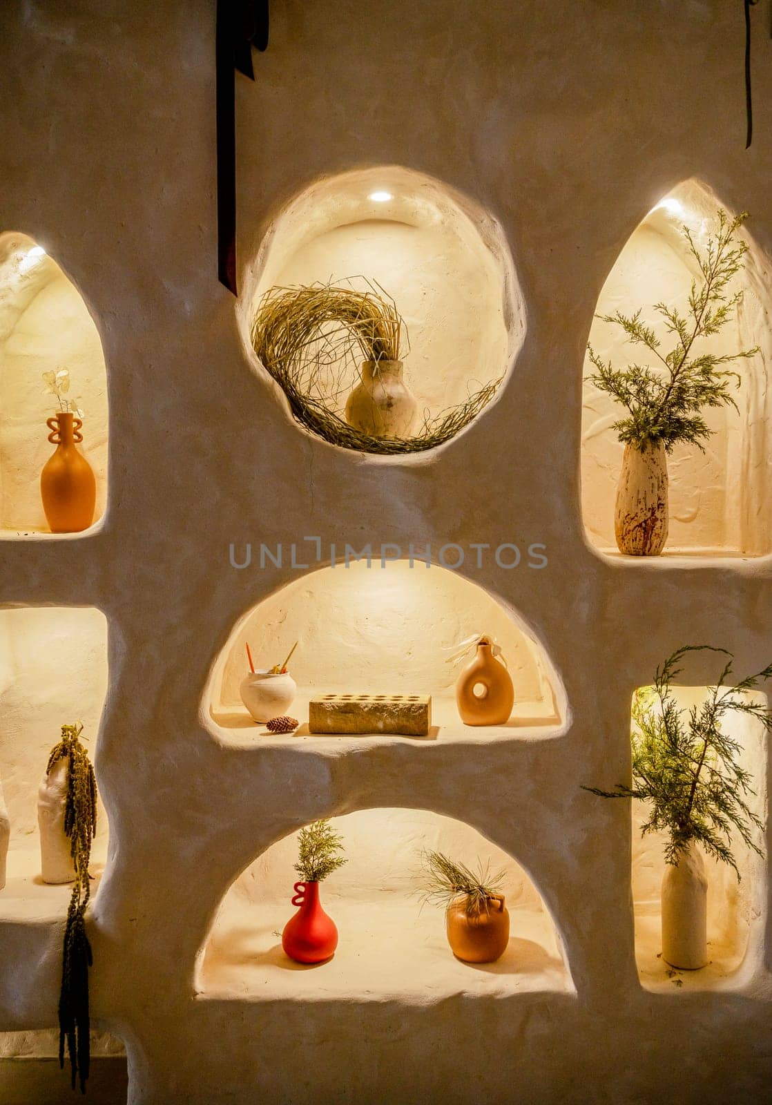 decorative wall with backlight and arches in which vases stand. beige wall in which vases with tree branches and flowers stand. arches are illuminated with warm light. handmade clay products