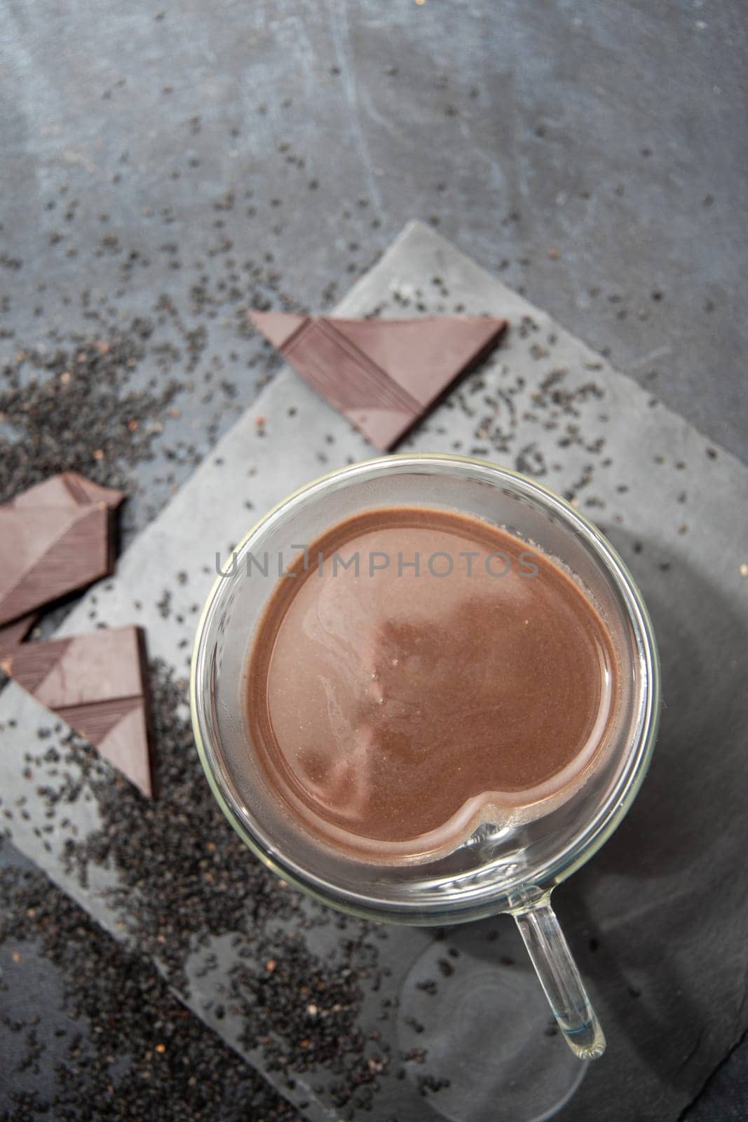 Glass cup of hot chocolate in heart shape on table surrounded by chocolate pieces and black chia seeds on grey textured background, Desert concept in color of the year 2025, mocha mousse, copy space