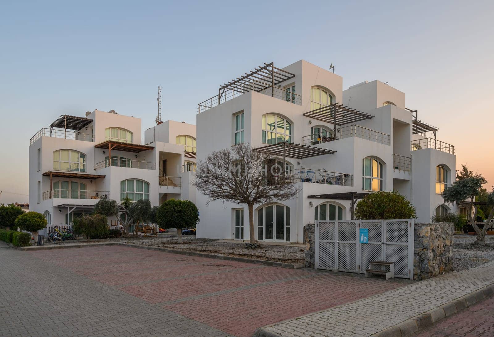 Charming white buildings adorned with balconies emerge amid lush greenery as twilight casts a warm glow.