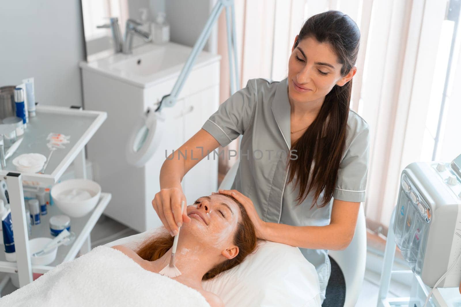 Professional cosmetology: Smiling massage therapist applying face mask to woman with brush at salon. From above of relaxed female customer is getting pampered in spa by beautician