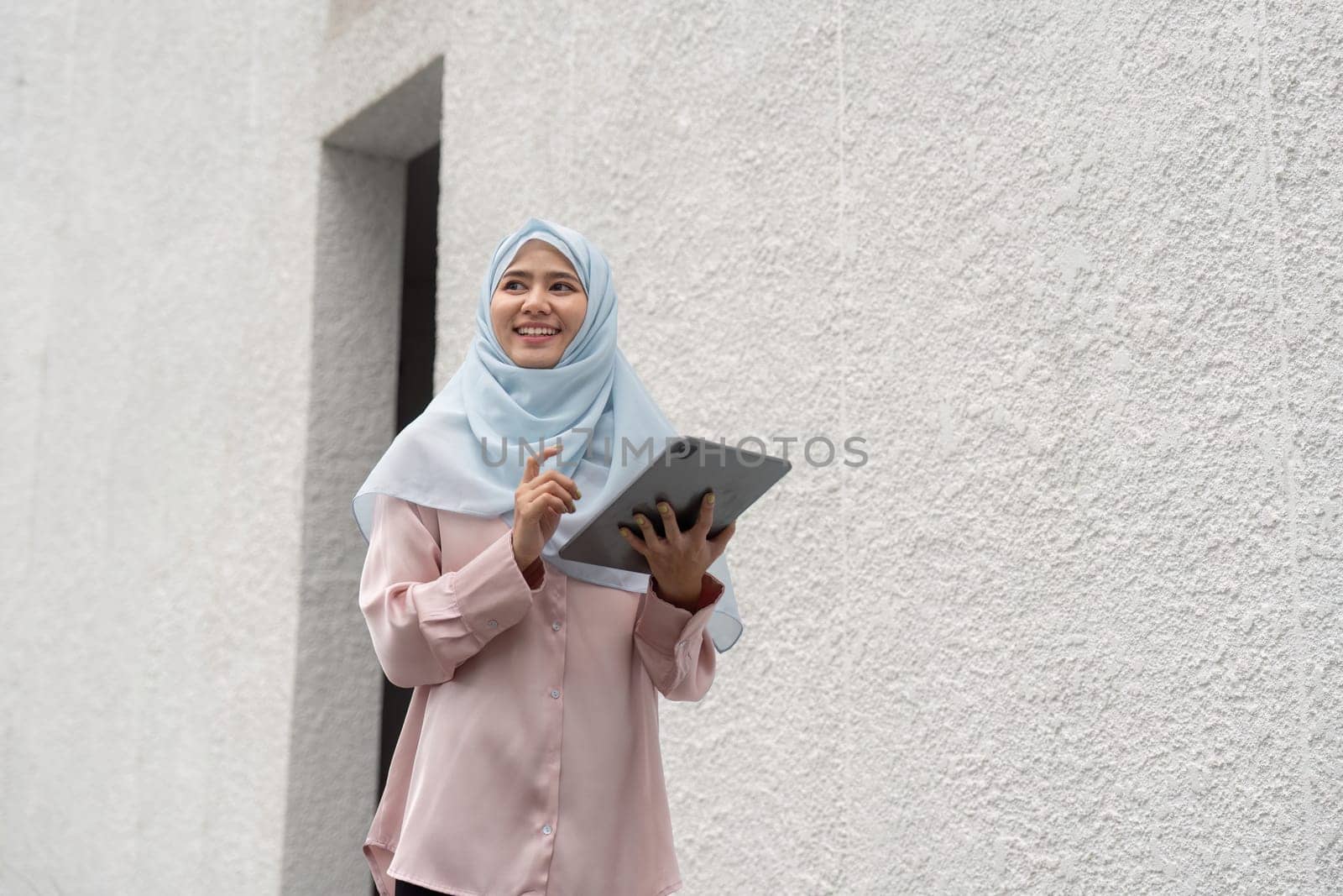 A smiling Muslim woman in a hijab interacts with her tablet, showcasing a blend of professionalism and technology in a contemporary business setting.