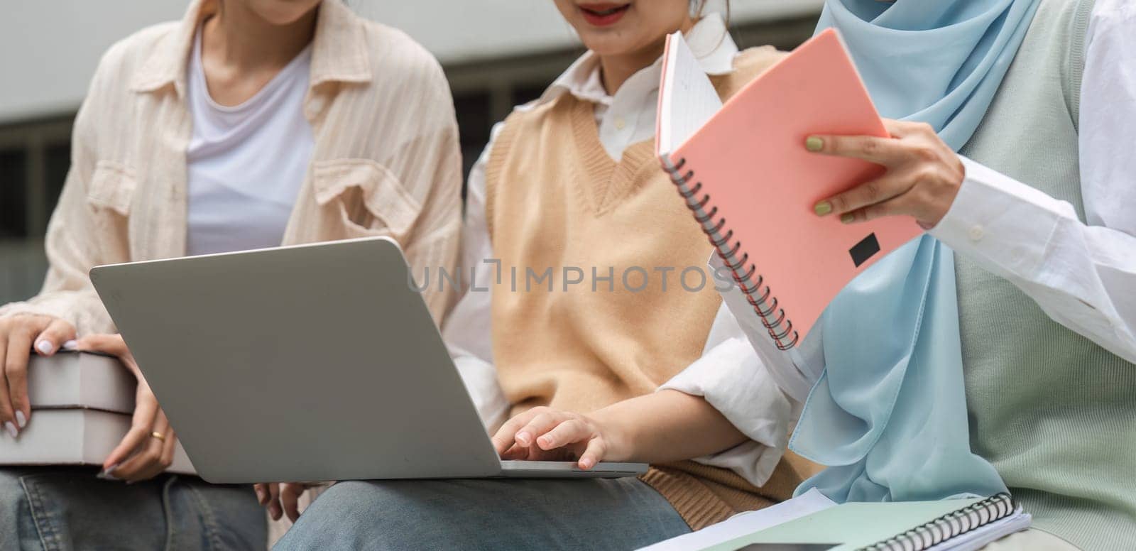 Close-up view of diverse students engaging in study with a laptop and notebooks while sitting outdoors at a university campus.