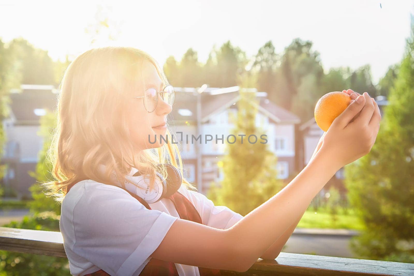 A girl examines a bright orange, basking in sunlight, surrounded by trees and homes during a serene afternoon.