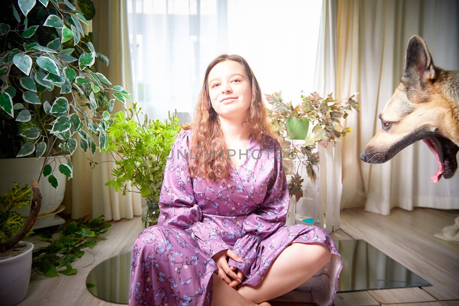 A young woman smiles while seated cross-legged in a bright room filled with greenery, as a dog playfully yells beside her.