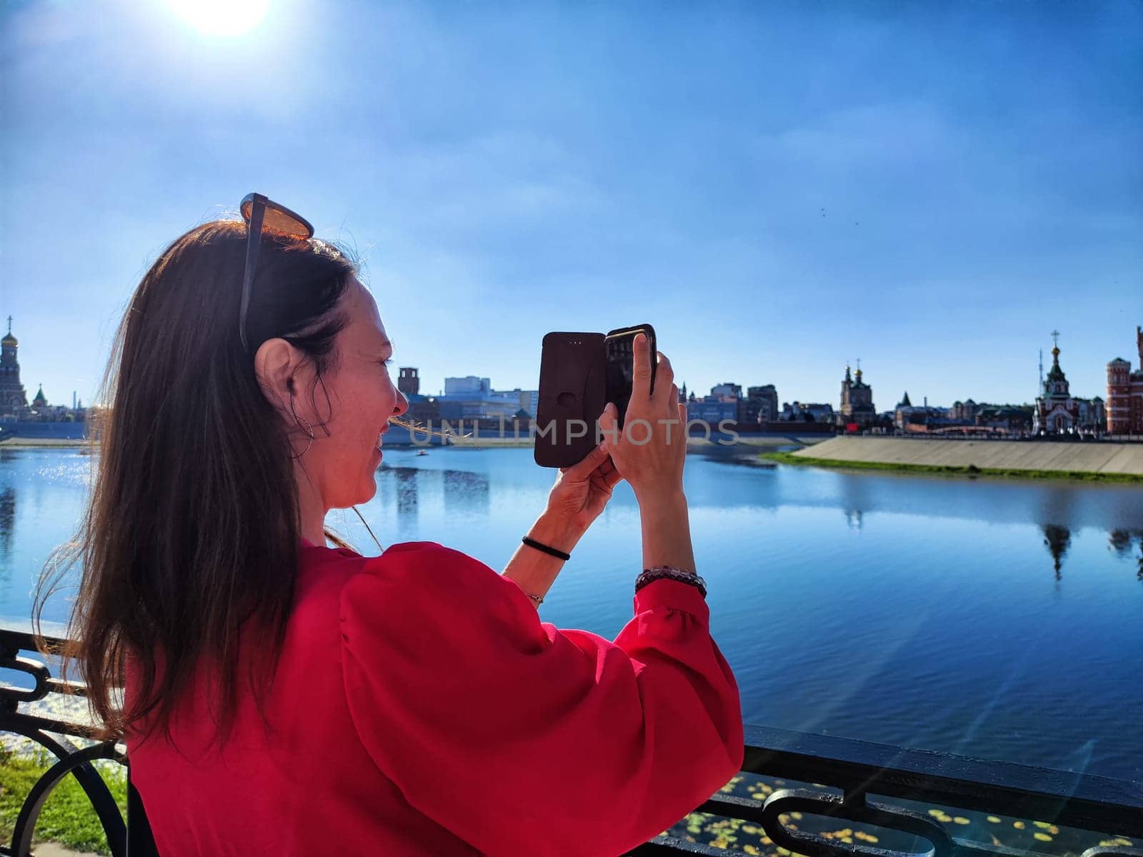 A woman in a red dress captures a beautiful skyline along the water under clear blue skies.
