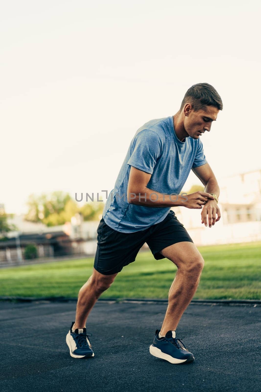 A man in a blue shirt and shorts checks his smartwatch while jogging on a track field. The scene is set during the afternoon with clear skies and green grass in the background.