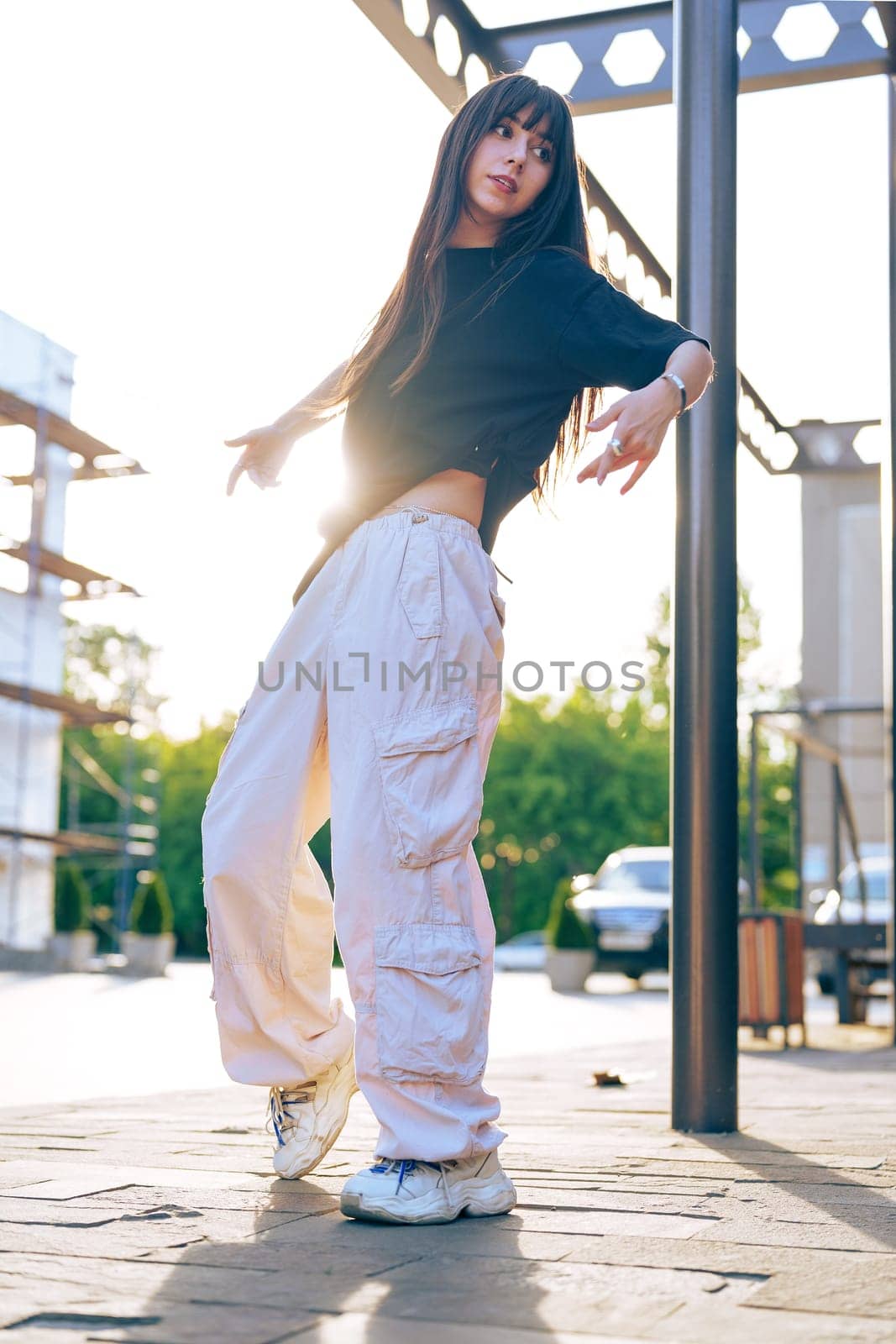 A young woman showcases her dance moves in an urban environment as the sun sets behind her. She wears loose, casual clothing and sneakers, exuding a vibrant energy.