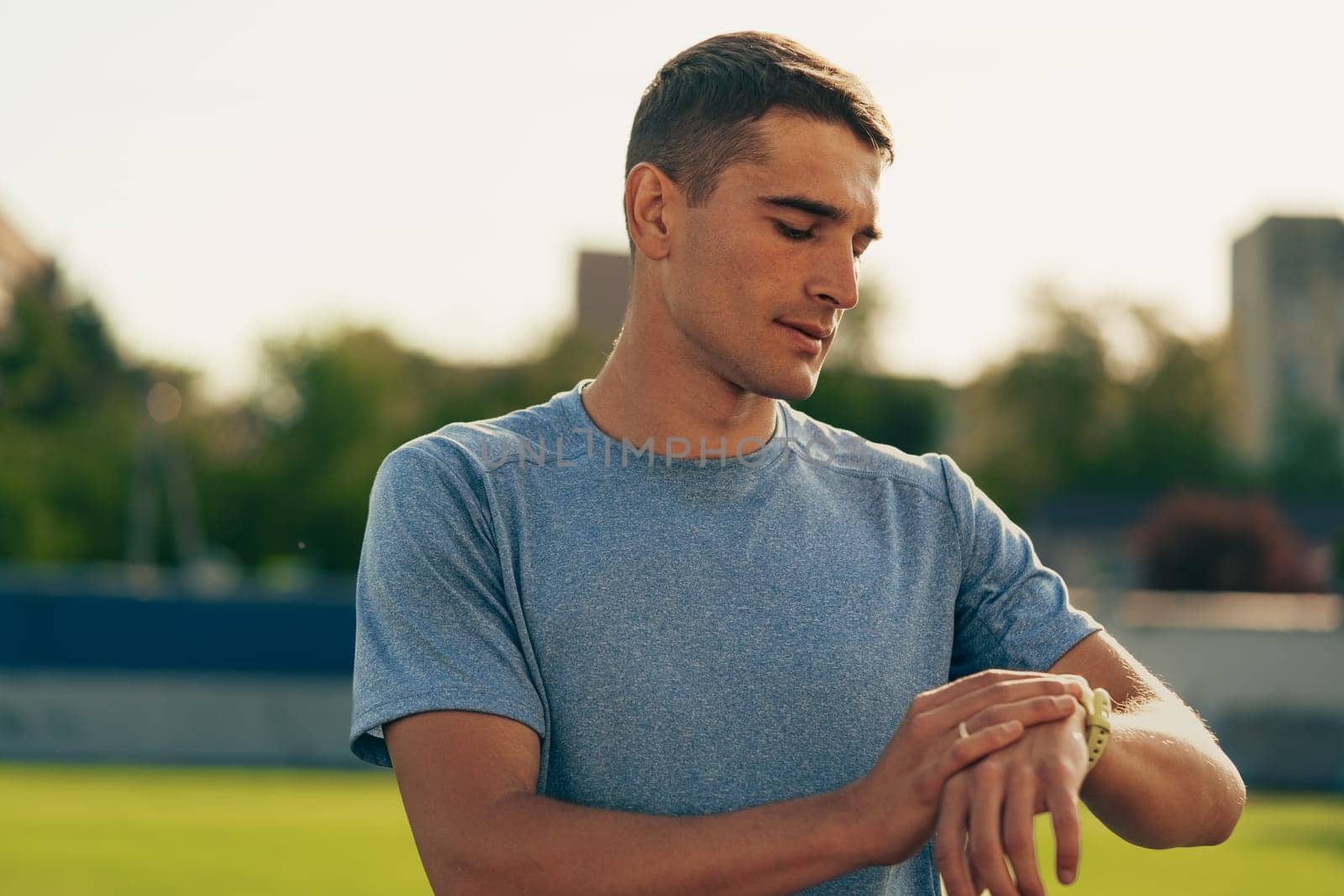 A young man wearing a blue athletic shirt is adjusting his watch while standing on an outdoor track. The sun is setting behind him, creating a warm glow around his focused expression.