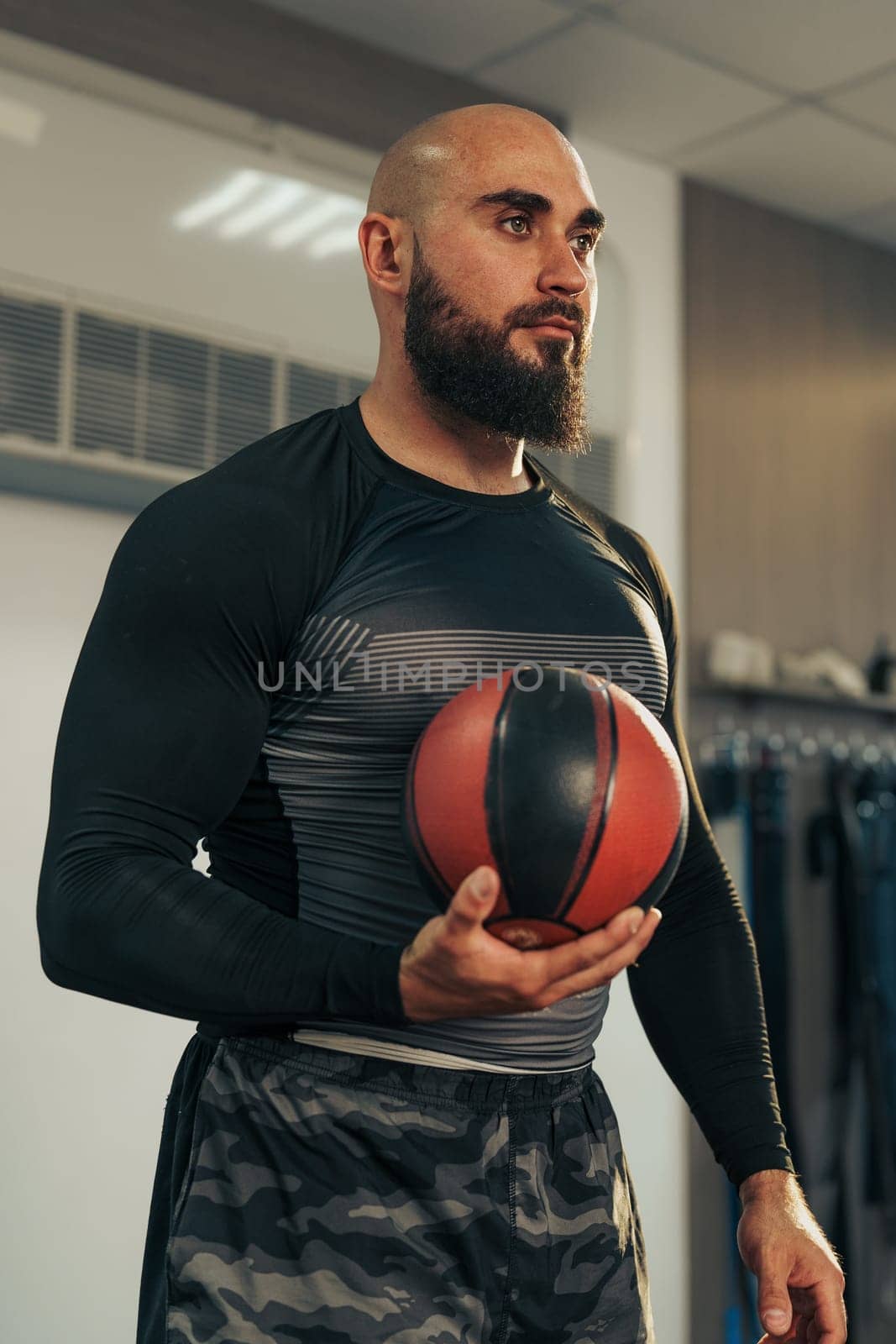 A strong male athlete stands in a gym, holding a basketball while focusing intently on his upcoming training session. He is dressed in a fitted workout outfit, reflecting dedication to sport.
