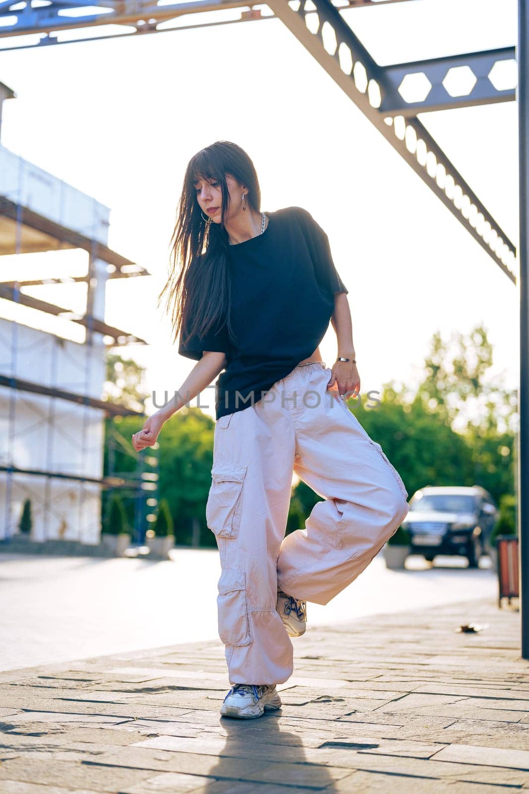 A young woman showcases her dance moves in an urban area, surrounded by construction and greenery, as the warm glow of golden hour enhances the atmosphere.