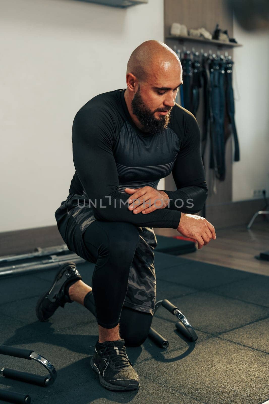 A man with a beard kneels on a gym mat, focused on his workout routine. He wears fitted training gear and demonstrates dedication to fitness in a well-equipped space.