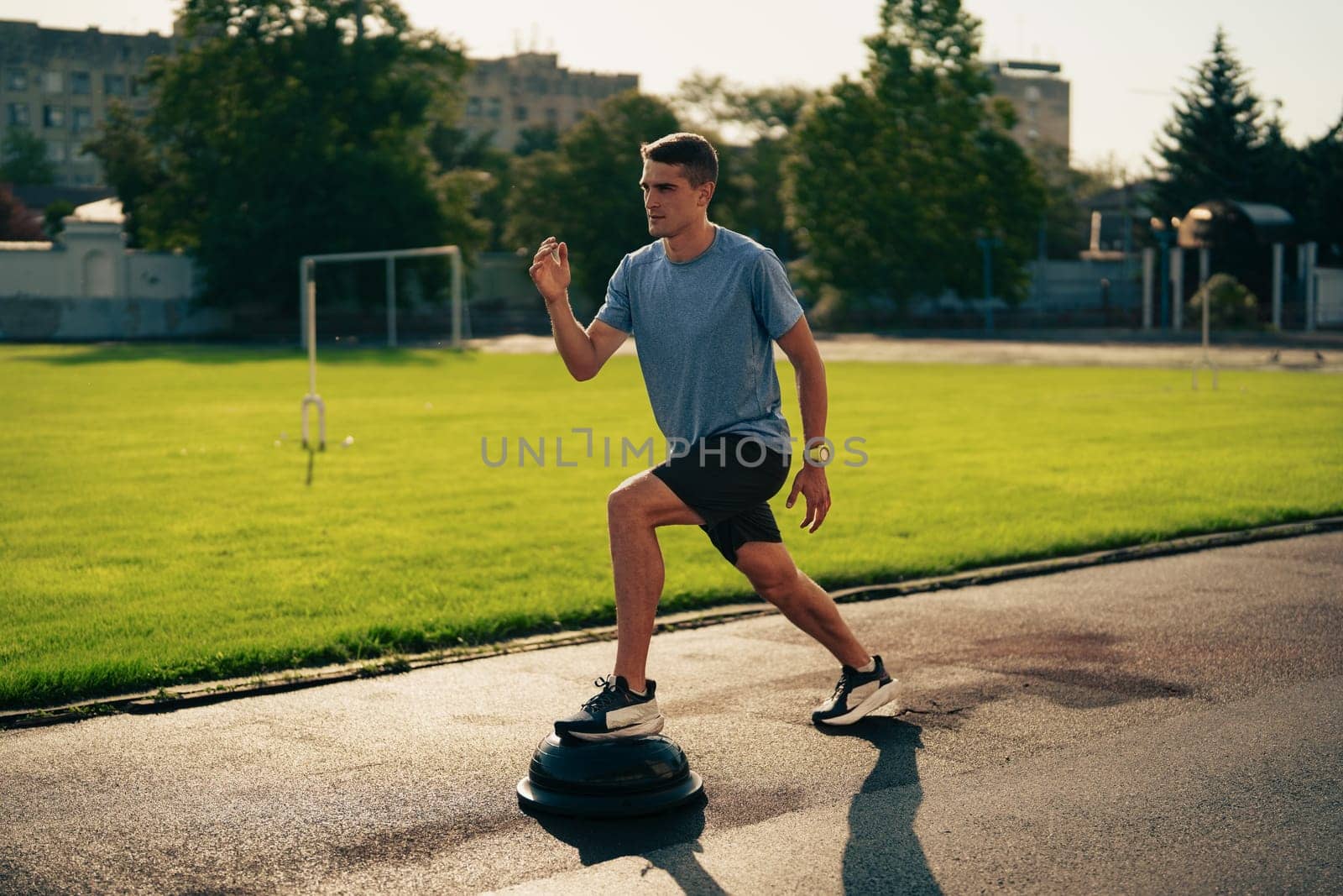 A young man is engaged in balance training on a stability disc at a city park during the late afternoon. The area is surrounded by grass and trees, providing a calm environment.