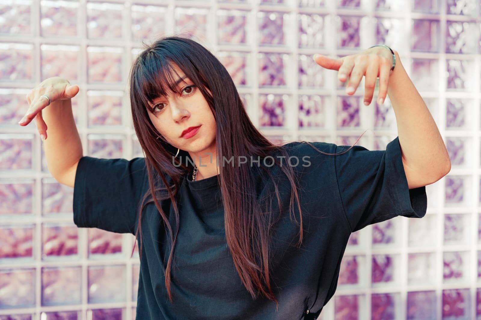 A young woman with long dark hair strikes a playful pose, showcasing her style against a translucent glass wall in a vibrant urban setting during daytime.