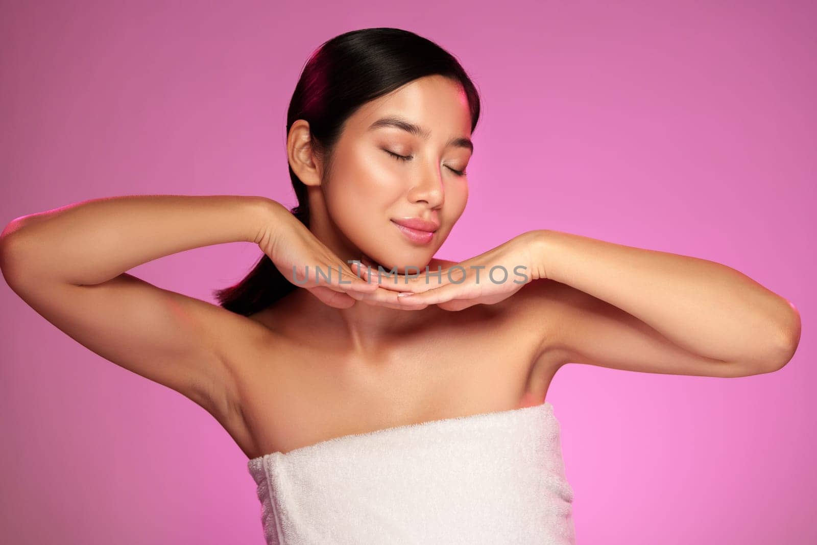 A model showcases her natural beauty with a radiant smile and relaxed pose while enjoying a beauty photoshoot against a vibrant pink background, emphasizing skin care.