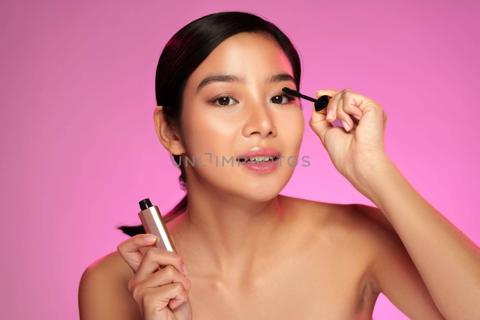 A woman showcases her makeup routine while applying mascara with precision. She smiles confidently, illuminated by soft lighting in a beauty studio, enhancing her natural features.