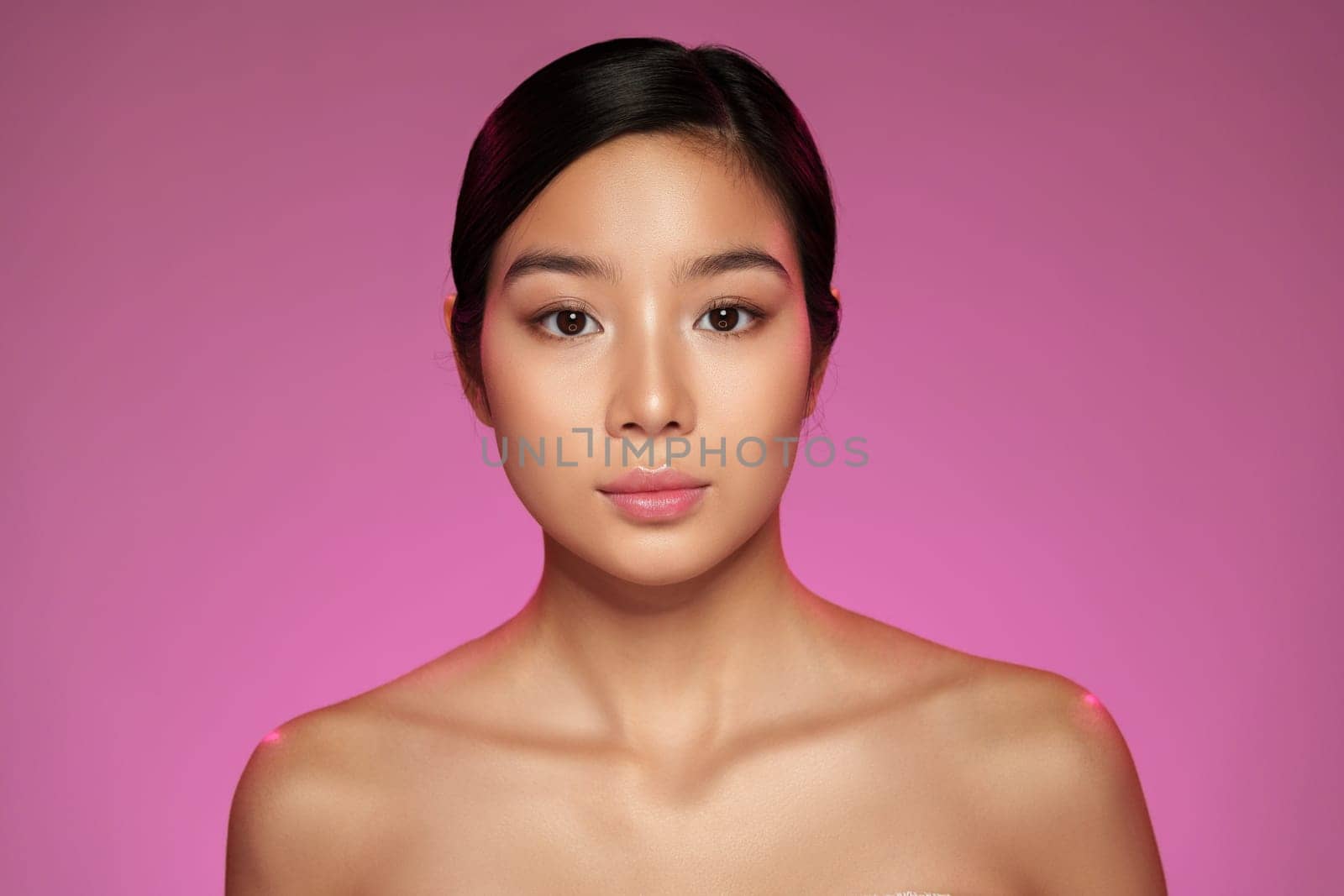 A woman stands confidently with a serene expression, showcasing natural beauty. The vibrant pink background adds a contemporary touch, enhancing her features.