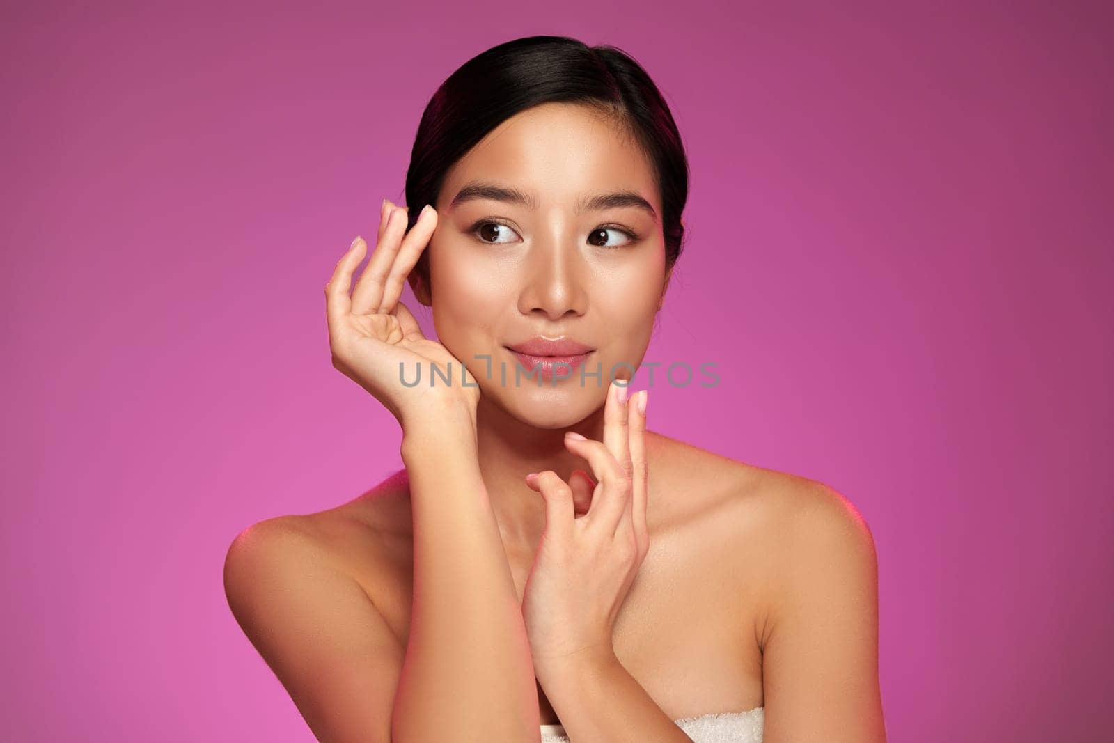A model gracefully poses with her hands near her face, displaying a serene expression. The vibrant pink backdrop emphasizes her features in a professional studio setting.