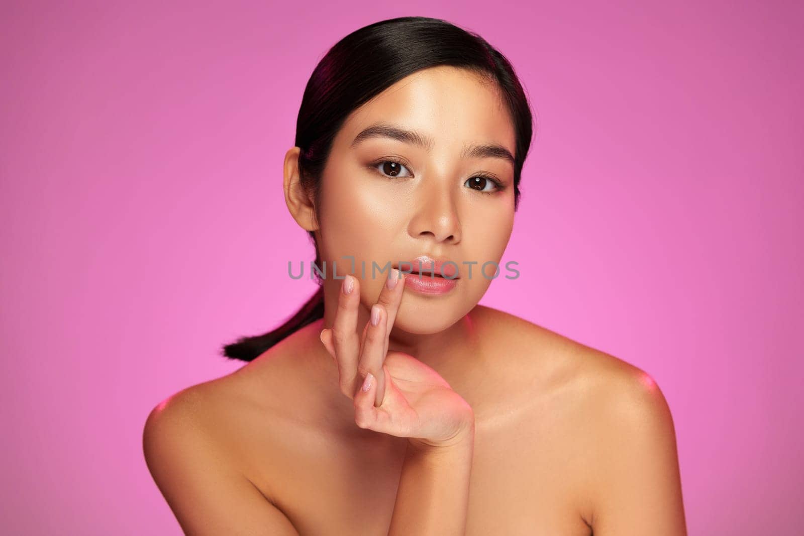 A model showcases her serene expression and flawless skin while gently touching her face against a vibrant pink background. The lighting emphasizes her features.