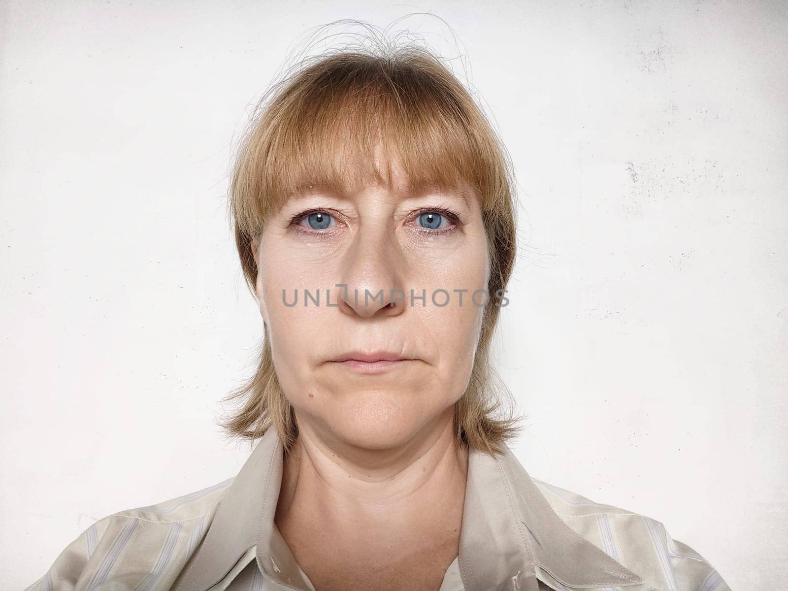 A middle-aged woman with blonde hair looks directly at the camera with a serious expression, reminiscent of a passport photo.