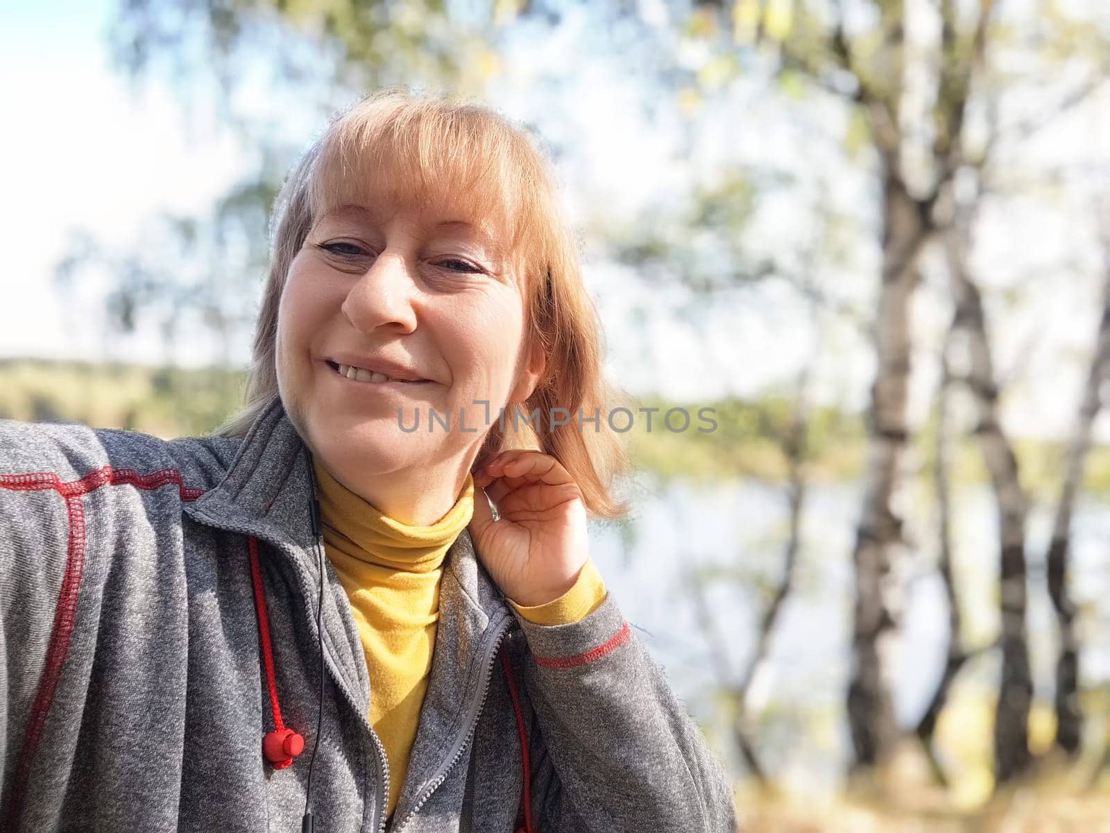 Blonde woman takes a cheerful selfie on a tranquil nature trip in autumn by a picturesque lake and vibrant fall foliage.