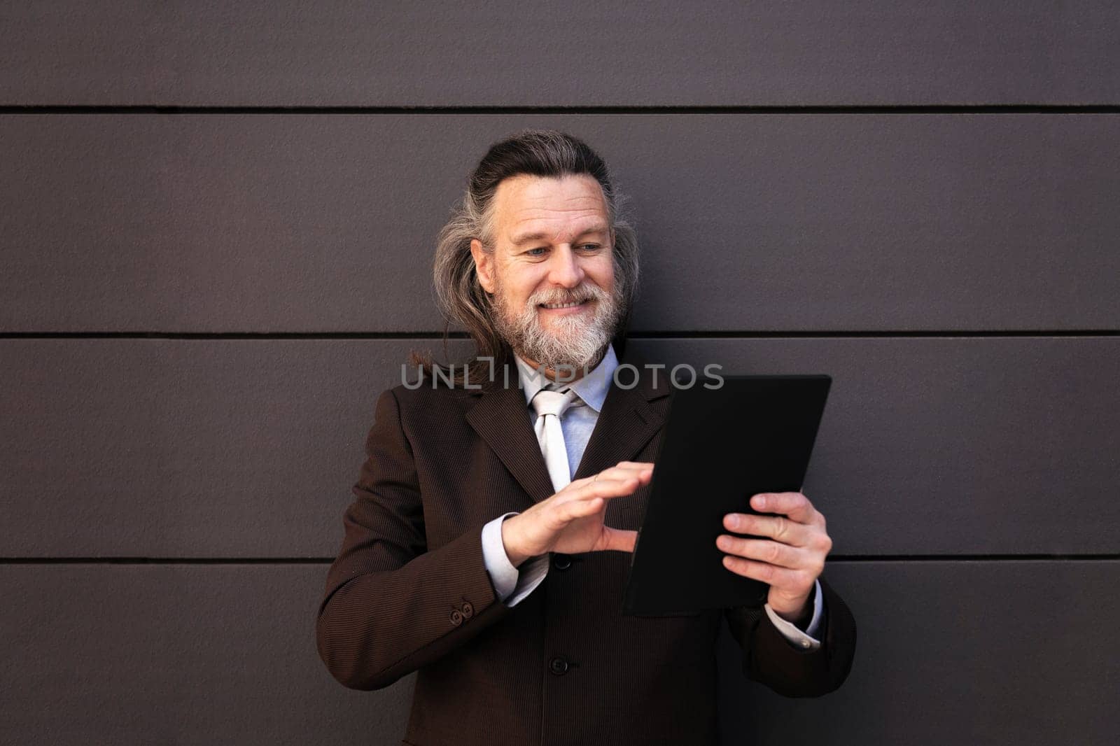 elegant mature man with beard in classy formal suit standing near gray wall and browsing internet on digital tablet, concept of middle age business man and technology of communication