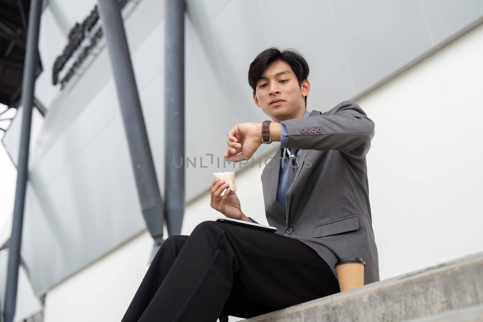 A young Asian businessman in a suit, checking his watch while eating a snack, balancing work and leisure in a contemporary outdoor environment.