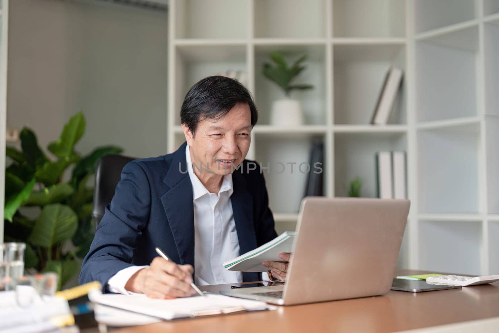 An elderly Asian businessman attentively reviewing documents and taking notes on his laptop in a contemporary office setting.