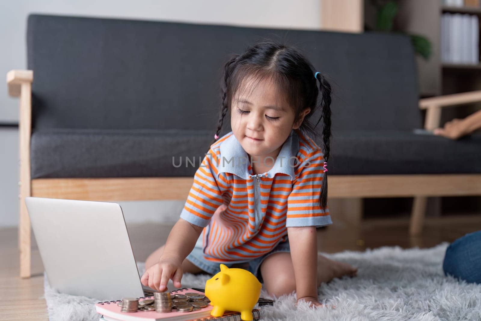 A young girl happily interacts with a laptop while exploring saving money concepts, surrounded by coins and a piggy bank in her warm living space.