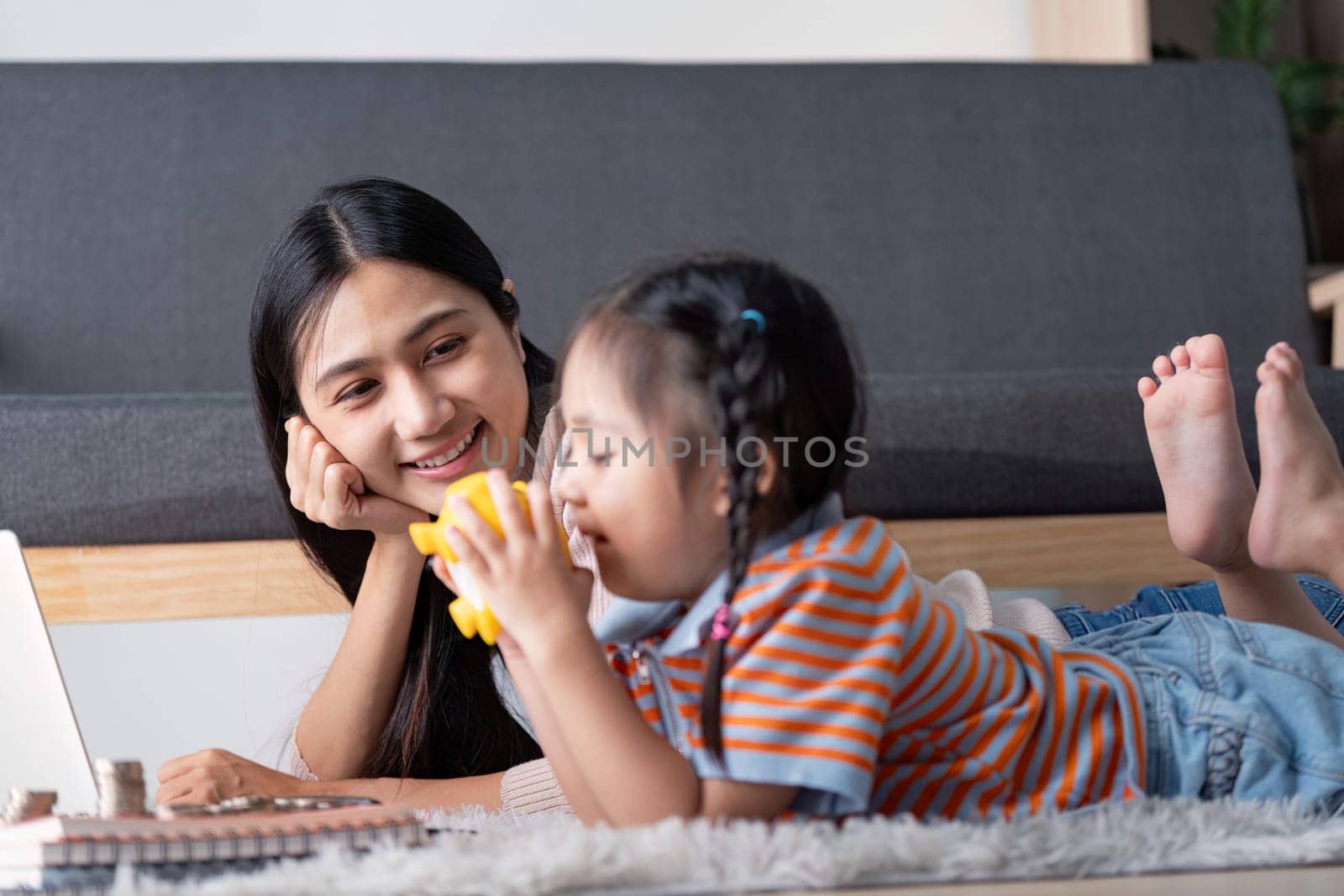 A diverse mother watches her daughter joyfully interact with a yellow piggy bank while they explore financial concepts together using a laptop at home.