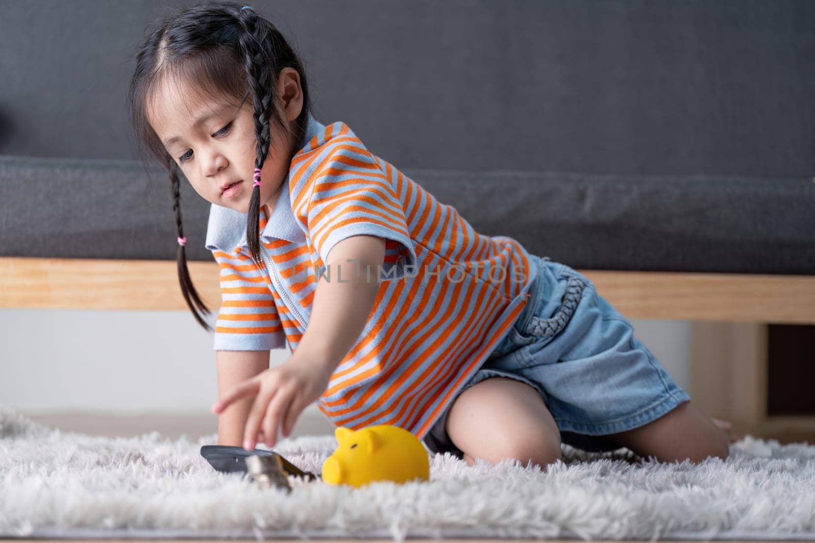 A young girl explores saving money by reaching for a yellow piggy bank and coins on the floor, promoting financial skills in a family setting.