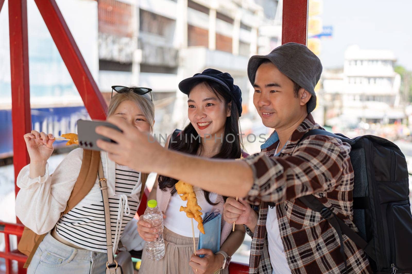 A group of young Asian friends happily taking a selfie with street food in hand, capturing joyful moments in a lively market atmosphere.