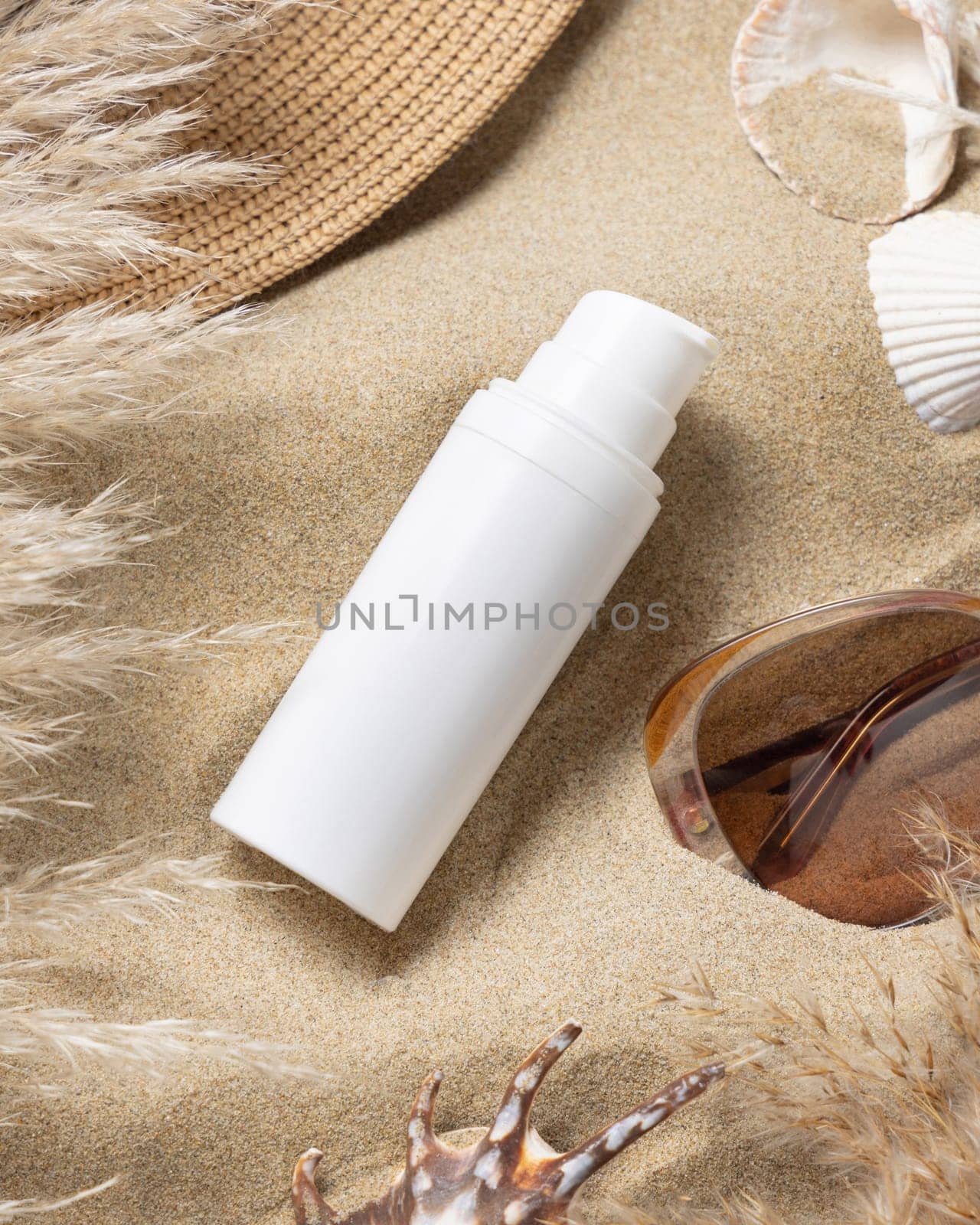 White blank one pump bottle, rattan hat, pampas grass and seashells on a sandy beach, top view. Sunscreen cosmetics mockup. Summer vacation with spf uv-protected sunbathing