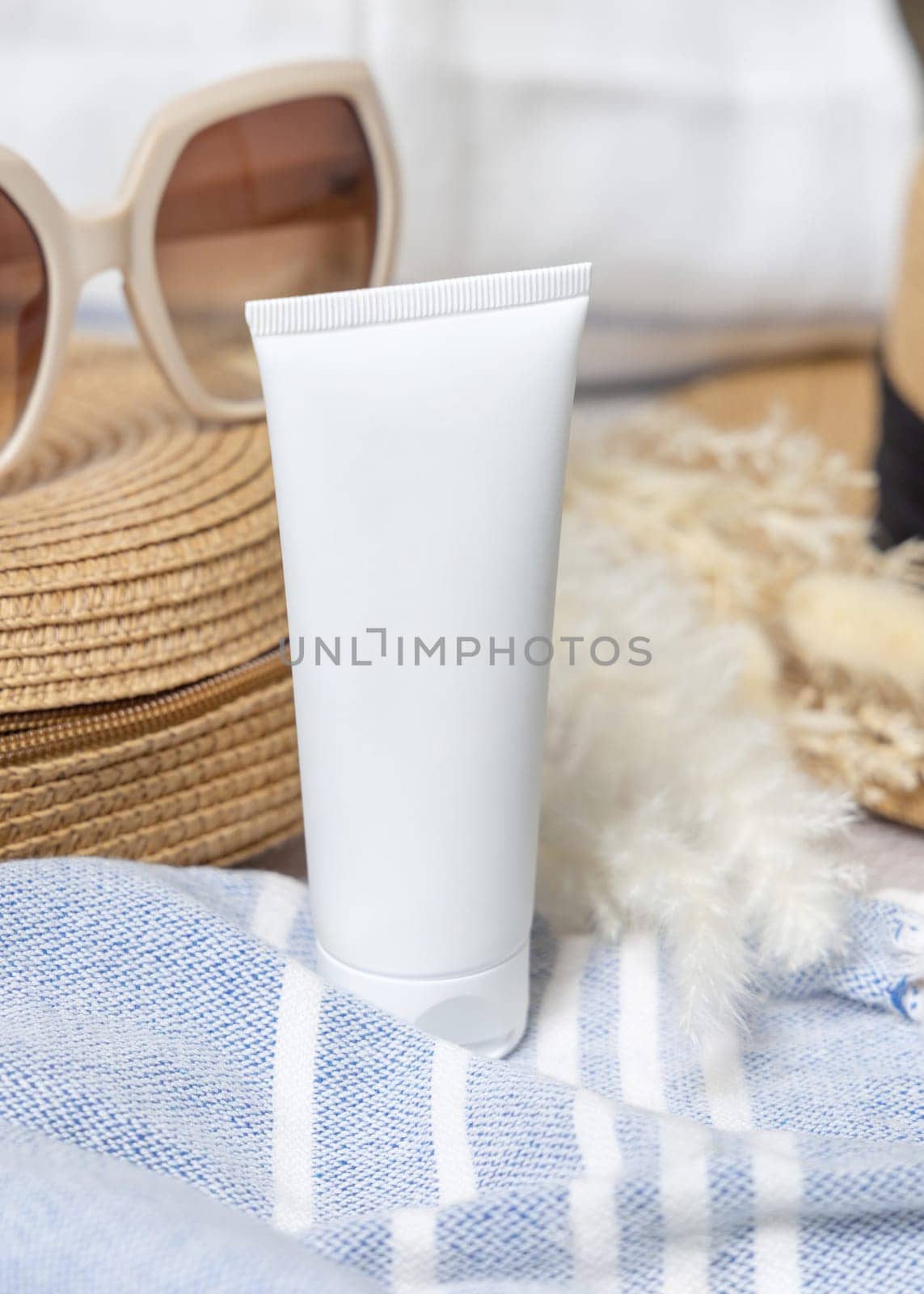 White blank squeeze cream tube on rattan bag near towel, dryed grass and sunglasses closeup. Sunscreen cosmetics mockup. Summer vacation with spf uv-protected sunbathing