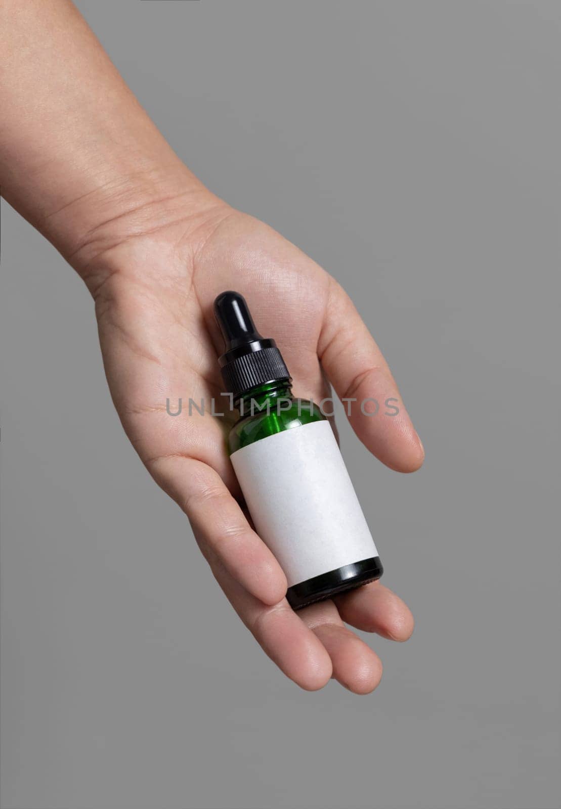 Female hand holding green dropper bottle with blank label against grey close up, cosmetic packaging mockup, copy space. Skincare beauty product - serum or essential oil
