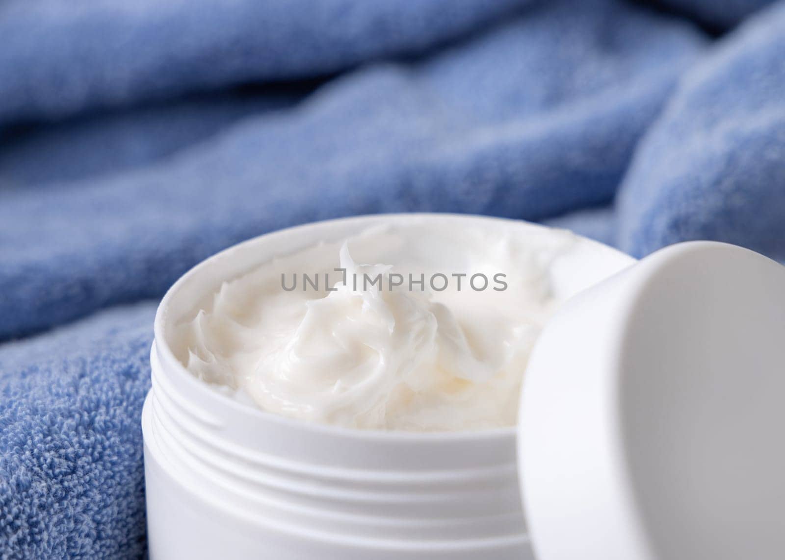 Cream jar on blue bath towel in bathroom, close up. Natural cosmetic product for facial care. Cream texture. Opened cosmetic container. Essence of Spa and Wellness