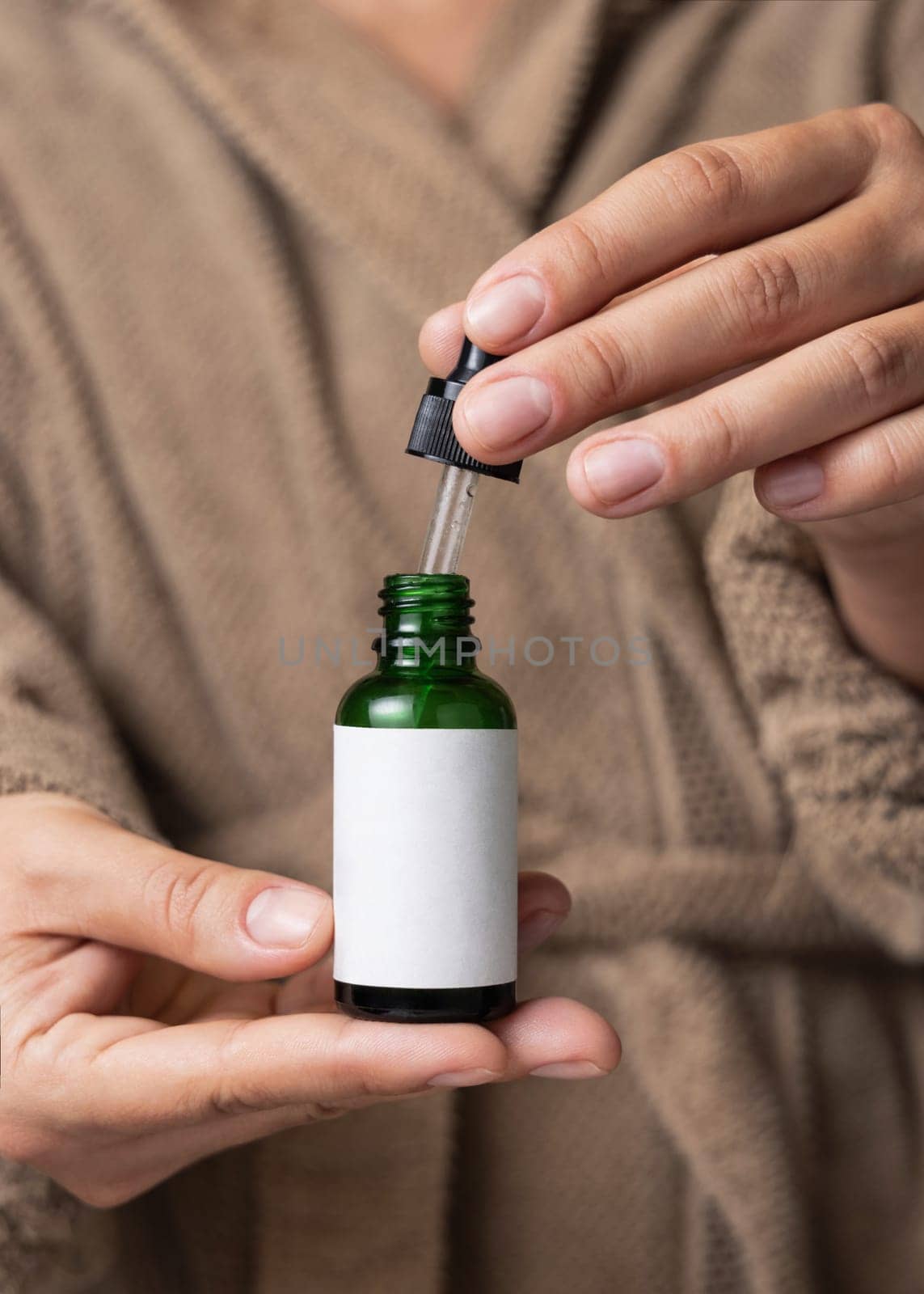 Female hands with natural manicure opening green cosmetic dropper bottle, label mockup. Close up of woman wearing light brown bathrobe using serum or essence