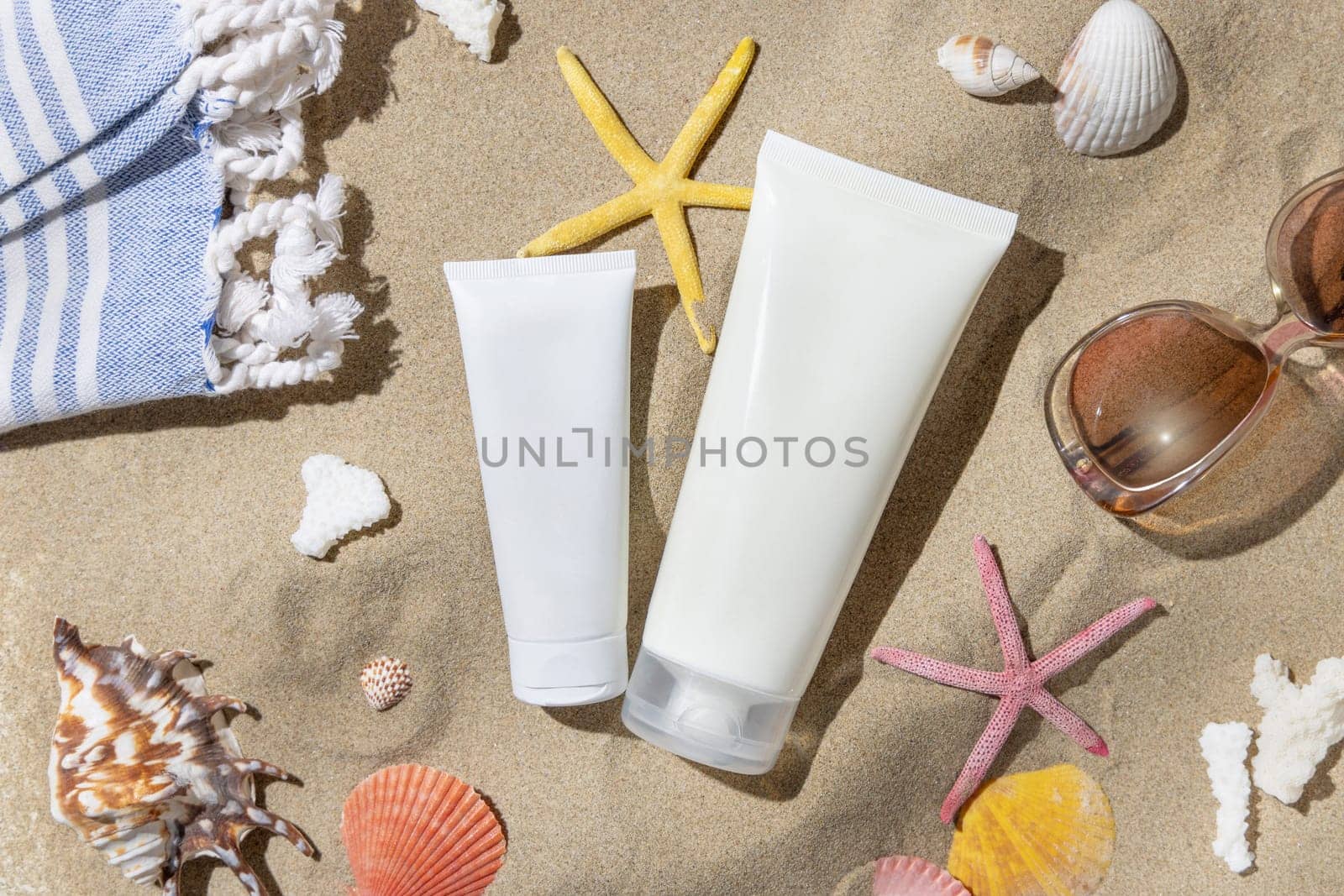 White blank cream tubes, starfishes and seashells on a sandy beach top view, hard shadow. Sunscreen cosmetics mockup. Summer vacation with spf uv-protected sunbathing