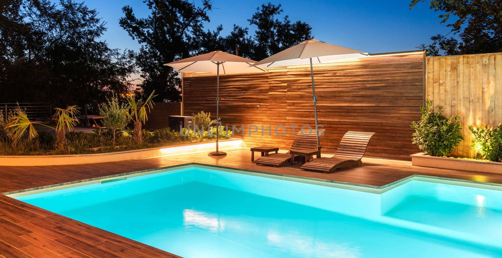 Modern backyard with illuminated swimming pool and lounge chairs under umbrellas. Luxury outdoor space surrounded by greenery and warm lighting
