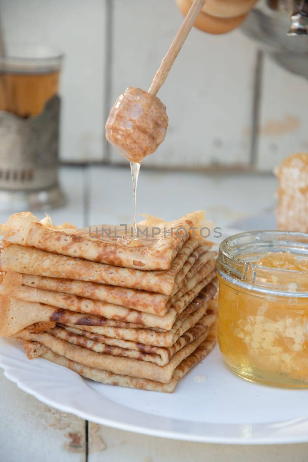 Drops of honey flow from a wooden spoon onto fresh pancakes, next to a cup of tea and a large samovar, a Russian tradition of celebrating Maslenitsa,