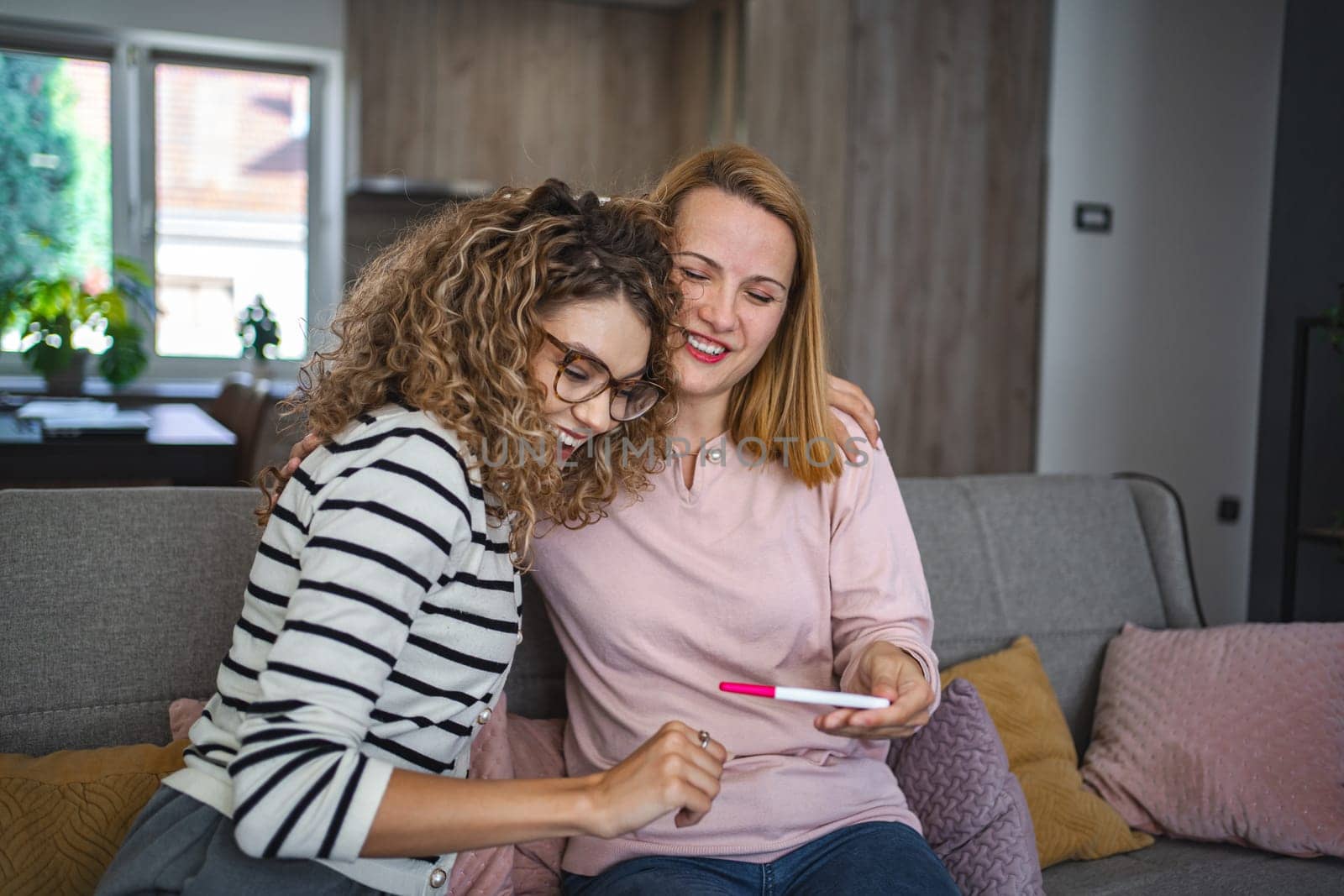 Two female friends are sitting on the couch in the living room and are excited after receiving the results of a pregnancy test