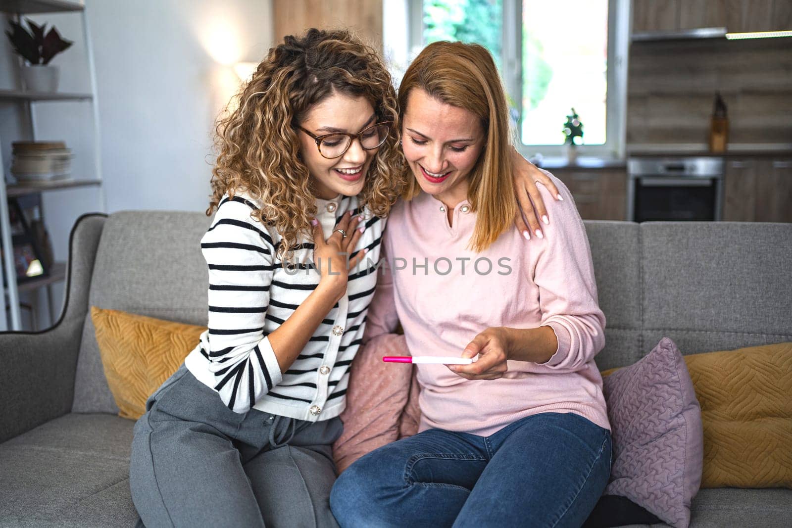 Two female friends are sitting on the couch in the living room, hugging and excited after getting their pregnancy test results.