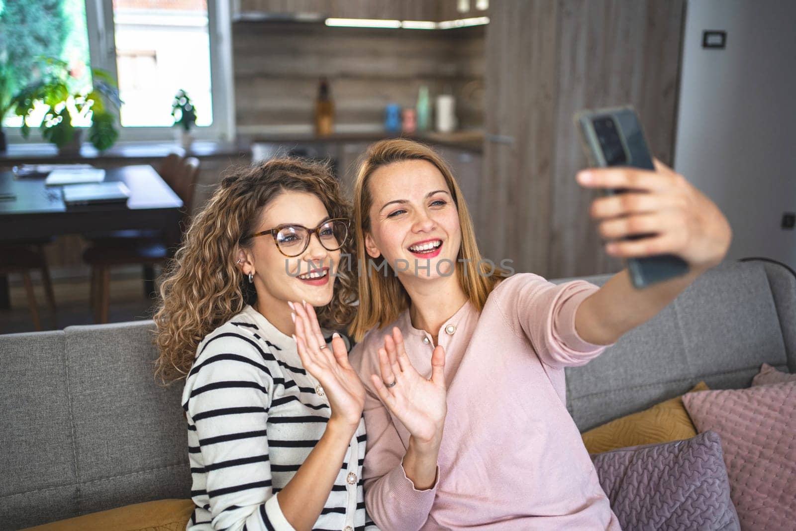 Two female friends are sitting on the couch in the living room and making a video call and sending greetings to the interlocutor.